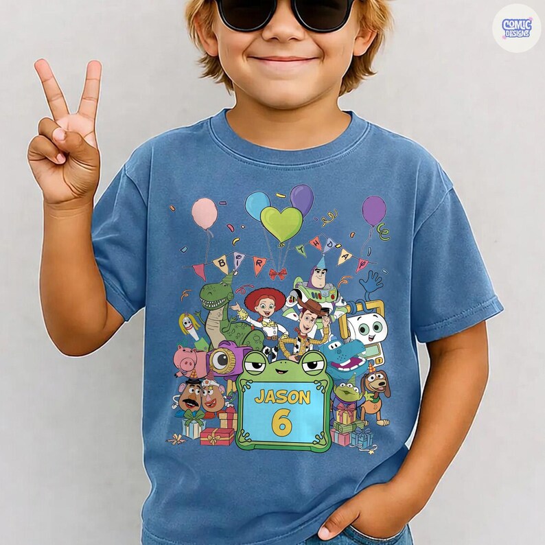 May include: A blue t-shirt featuring a colorful graphic print of various animated toy characters, including a cowboy, space ranger, and dinosaur. Above the characters are balloons and a banner. A green frame contains the text JASON and the number 6.