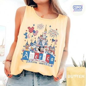 May include: A pale yellow sleeveless vest top featuring a graphic print of a castle, fireworks, and cartoon characters in red, white, and blue. The text reads America, 250 Years of Freedom, and 1776-2026, with a small Comic Designs logo in the corner.
