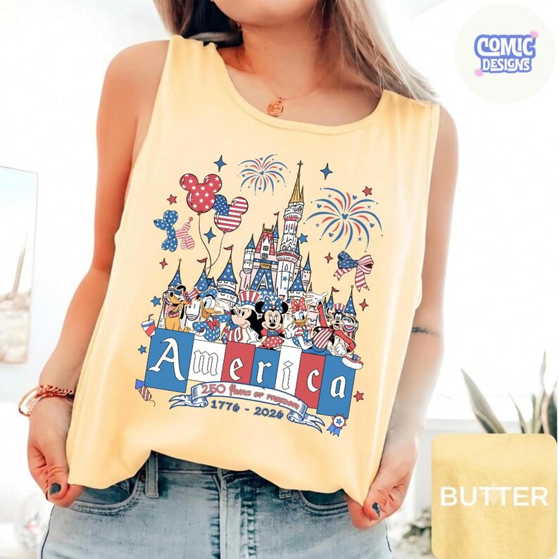 May include: A pale yellow sleeveless vest top featuring a graphic print of a castle, fireworks, and cartoon characters in red, white, and blue. The text reads America, 250 Years of Freedom, and 1776-2026, with a small Comic Designs logo in the corner.