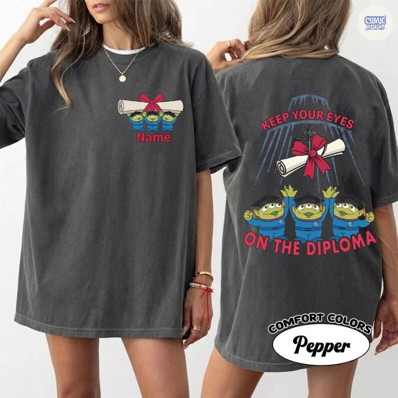May include: Dark grey t-shirt featuring a cartoon design of three green aliens holding a diploma with a red bow. The front of the shirt has the word "Name" below the aliens. The back of the shirt says "Keep Your Eyes On The Diploma".