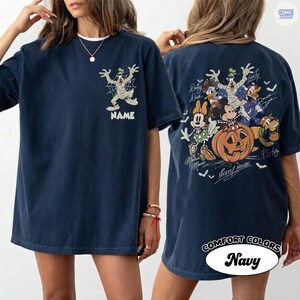 May include: A navy blue t-shirt featuring a graphic of Goofy dressed as a mummy on the front chest above the word NAME. The back displays a large Halloween-themed illustration of Mickey Mouse, Minnie Mouse, Donald Duck, Daisy Duck, Goofy, and Pluto.