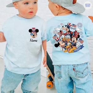 May include: Light blue t-shirt featuring Mickey Mouse in patriotic attire with the text "Name". The back of the shirt displays Mickey, Minnie, Donald, Goofy, and Pluto with American flags and fireworks. The shirt is part of the "America 250th USA Anniversary" collection.