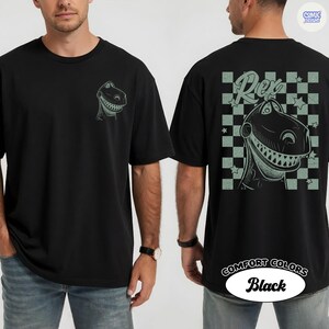 Puede incluir: Camiseta negra con un dise&ntilde;o de dinosaurio de dibujos animados, "Rex", en la parte delantera y trasera. El dise&ntilde;o trasero incluye un patr&oacute;n de cuadros y el nombre "Rex". La camiseta est&aacute; etiquetada como "Comfort Colors".
