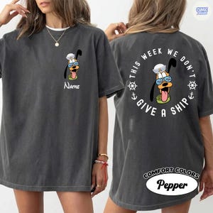 May include: Dark grey t-shirt featuring a cartoon Pluto character wearing a sailor hat and glasses. The front of the shirt has the character's face and the name "Name". The back of the shirt has the text "This week we don't give a ship" around the character.