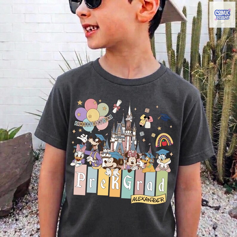 May include: Dark gray t-shirt featuring a colorful graphic of Disney characters, a castle, balloons, and the text "Congratulations" and "Prek Grad Alexander". The design includes a rainbow and graduation caps.