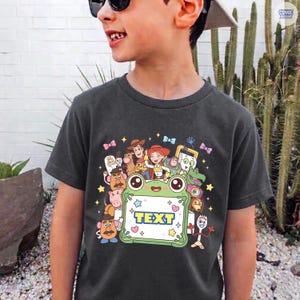 May include: A dark gray t-shirt featuring a colorful graphic print of various animated characters surrounding a central light green frog-shaped frame with the word TEXT printed in blue letters inside, accented by small stars and hearts.
