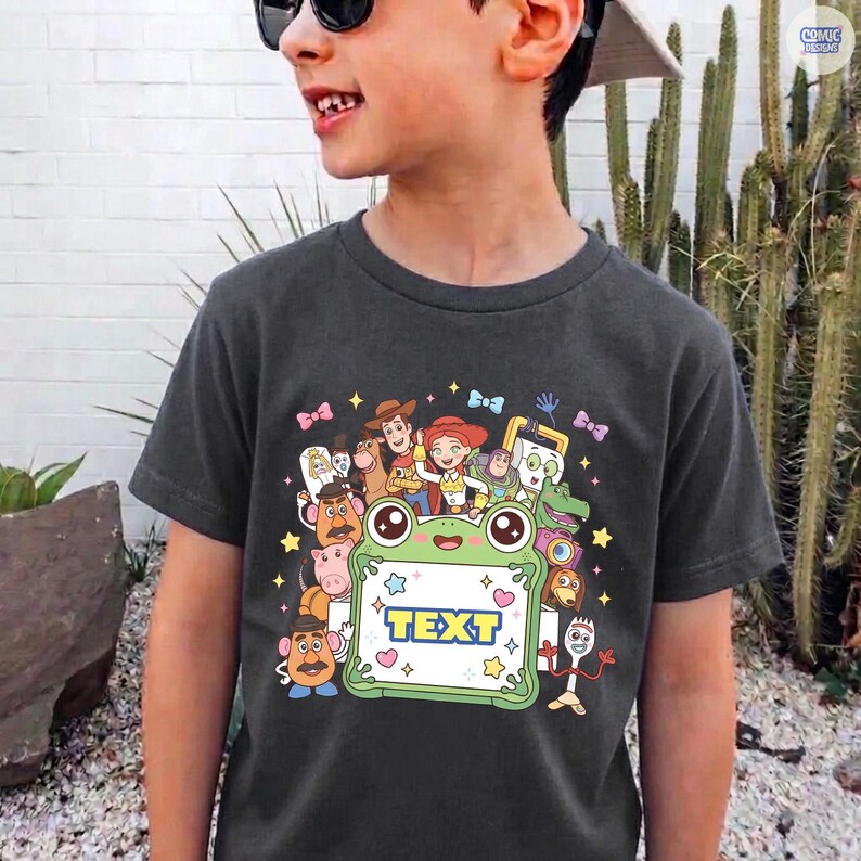 May include: A dark gray t-shirt featuring a colorful graphic print of various animated characters surrounding a central light green frog-shaped frame with the word TEXT printed in blue letters inside, accented by small stars and hearts.