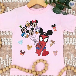 Peut inclure: T-shirt rose clair avec un personnage de Spider-Man tenant des ballons en forme de personnages Disney. Graphiques Disney suppl&eacute;mentaires, dont Mickey Mouse, une glace et une tasse de caf&eacute;. Le t-shirt est sur un cintre en bois.