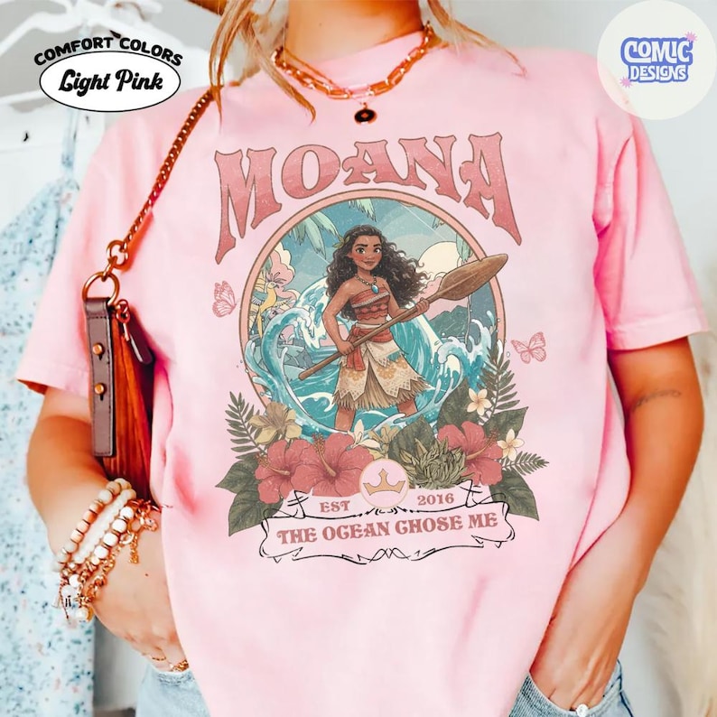 May include: A light pink t-shirt featuring a graphic of a character holding a wooden oar in front of ocean waves. The design includes tropical flowers, butterflies, and the text MOANA and THE OGEAN GHOSE ME. A gold chain necklace is worn.