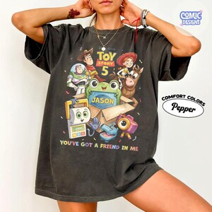 May include: A charcoal gray t-shirt featuring a Toy Story 5 graphic with characters including Woody, Buzz Lightyear, Jessie, and Bullseye. A green frog holds a box labeled Jason. The text reads Toy Story 5 and You've Got A Friend In Me.