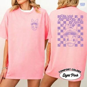 May include: Pale pink t-shirt with a purple Daisy Duck design on the front and back. The back features the text "Daisy Duck" above a chequered pattern with stars. The shirt is labelled "Comfort Colors" and "Light Pink".