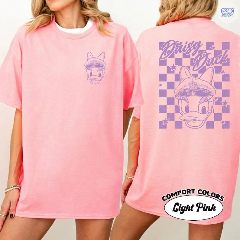 May include: Pale pink t-shirt with a purple Daisy Duck design on the front and back. The back features the text "Daisy Duck" above a chequered pattern with stars. The shirt is labelled "Comfort Colors" and "Light Pink".