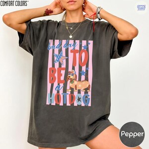 May include: Pepper-colored Comfort Colors t-shirt with a graphic that reads "bad day to be a hotdog." The graphic features a dachshund wearing sunglasses and a hotdog costume, set against pink and blue stripes and stars.