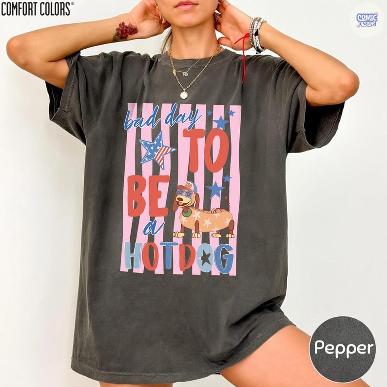 May include: Pepper-colored Comfort Colors t-shirt with a graphic that reads "bad day to be a hotdog." The graphic features a dachshund wearing sunglasses and a hotdog costume, set against pink and blue stripes and stars.