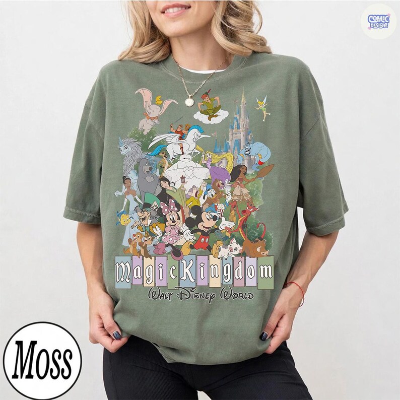 Puede incluir: Camiseta verde oliva con un gr&aacute;fico colorido de varios personajes de Disney, incluyendo Mickey Mouse, Peter Pan y el castillo de Cenicienta. El texto "Magic Kingdom Walt Disney World" se muestra debajo.