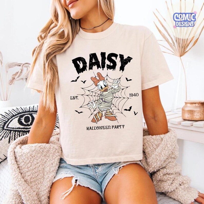 May include: A cream-colored t-shirt featuring a graphic of Daisy Duck wrapped in mummy bandages standing in a spider web. The text reads DAISY, EST. 1940, and HALLOWEEN PARTY, accompanied by small black bat illustrations surrounding the central character.