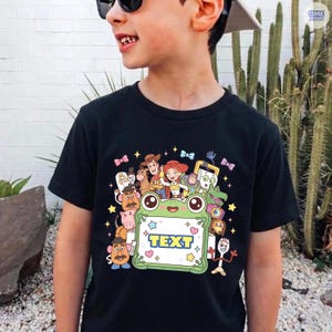 May include: A black t-shirt featuring a colorful graphic print of various animated characters surrounding a central light green frog-shaped frame with the word TEXT printed in bold blue letters, accented by small stars and bows.