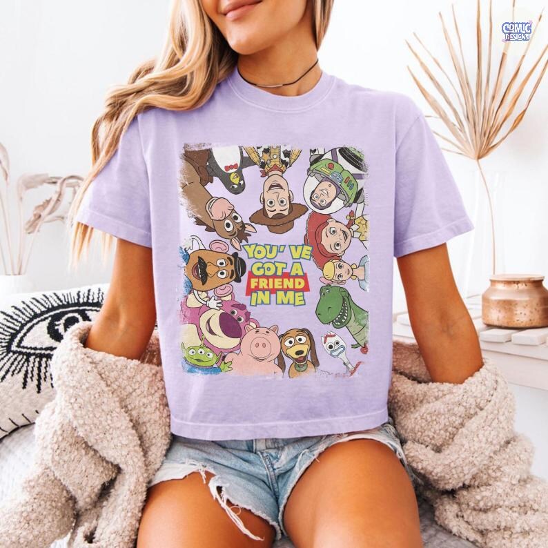 May include: A light purple t-shirt featuring a collage of characters from the animated movie Toy Story. The shirt includes the text "YOU'VE GOT A FRIEND IN ME" in a red and white design.