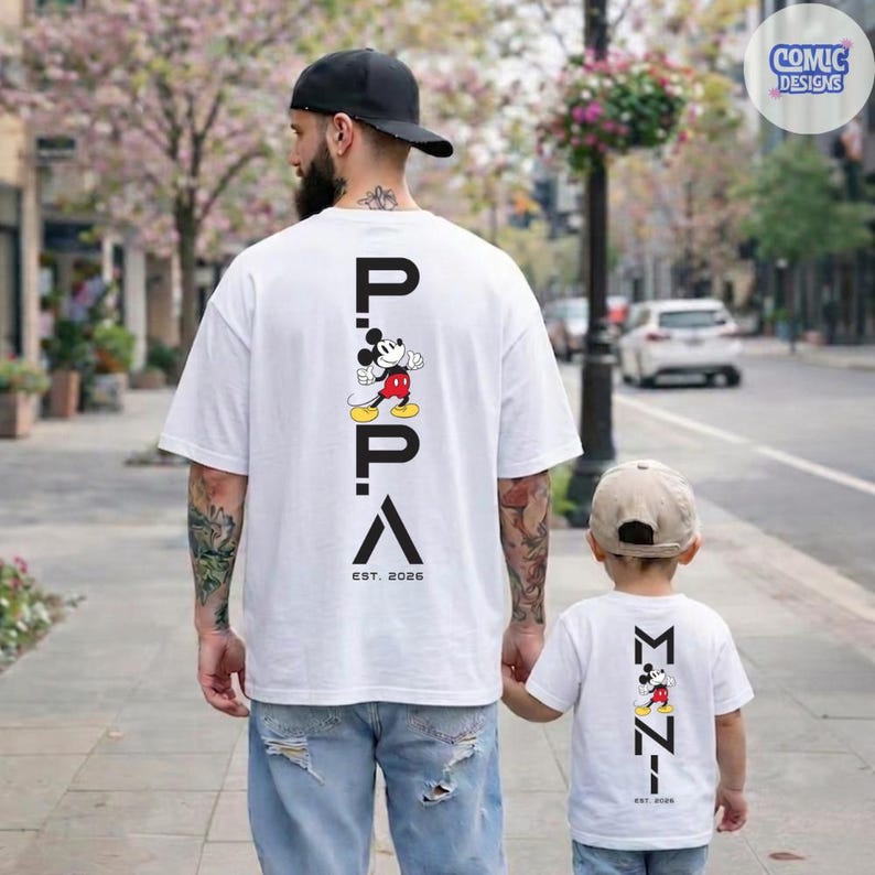 May include: White t-shirts with a Mickey Mouse graphic and the text "PAPA" and "EST. 2026" on the back. A father and child are wearing matching shirts. The father is wearing a black cap and blue jeans.