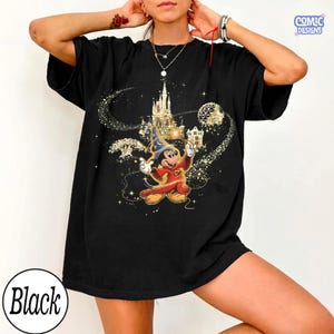 May include: Black t-shirt featuring a graphic of Mickey Mouse as a sorcerer in red and gold attire, with a castle and sparkling accents. The text "COMIC DESIGNS" is in the upper right corner, and "Black" is in a circle at the bottom.