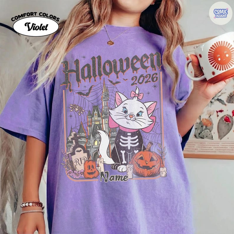May include: A lilac Comfort Colors t-shirt with a Halloween 2026 graphic. The design includes a cartoon cat in a skeleton costume, a haunted castle, pumpkins, and the word "Name". The shirt has a relaxed fit.
