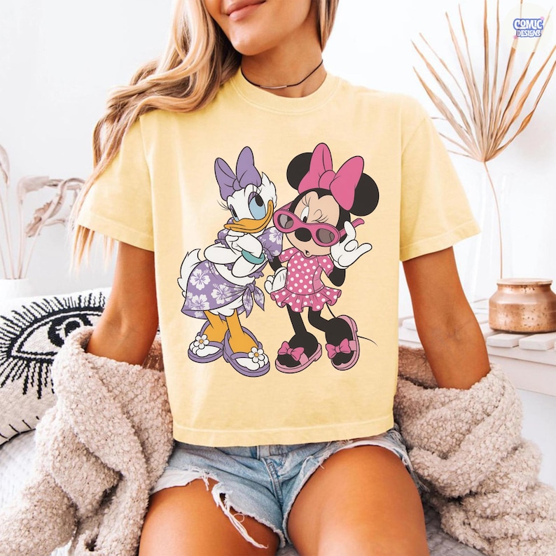 Puede incluir: Una camiseta de color amarillo p&aacute;lido con un estampado gr&aacute;fico de Daisy Duck y Minnie Mouse. Daisy lleva un vestido morado con flores y un lazo, mientras que Minnie lleva un vestido rosa de lunares, un lazo rosa y gafas de sol rosas. Ambas llevan sandalias.