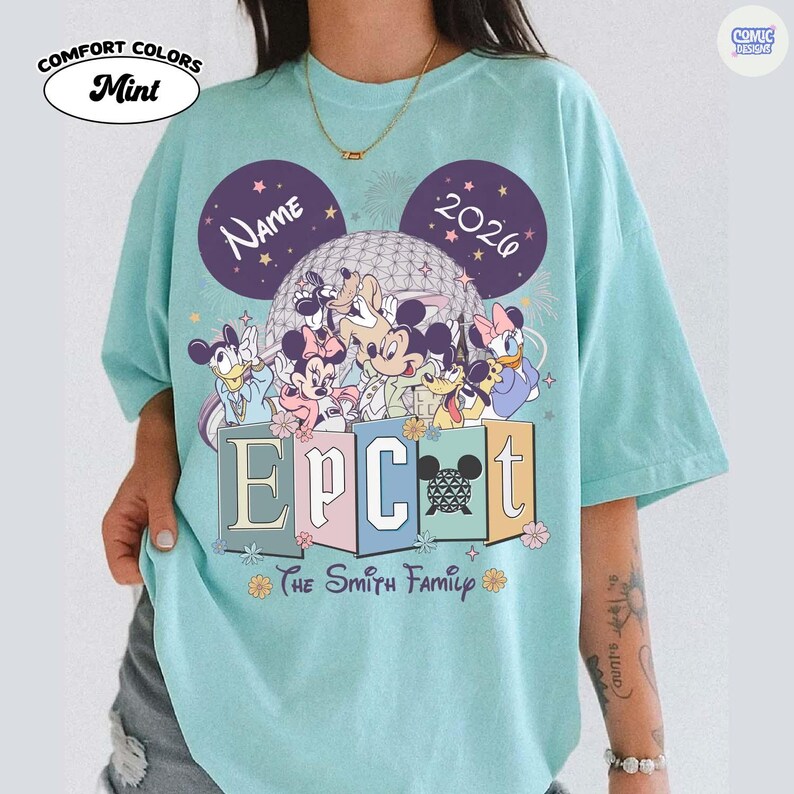 May include: Mint green Comfort Colors t-shirt featuring a Disney theme. The design includes Mickey Mouse ears, the text "Name 2026", and the word "EPCOT" with Disney characters. The text "The Smith Family" is also present.