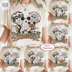 Peut inclure: Un t-shirt couleur cr&egrave;me avec une impression graphique repr&eacute;sentant un personnage fant&ocirc;me portant des oreilles de souris, tenant une citrouille devant une toile d'araign&eacute;e et des branches d'arbre. Le texte BOO et HALLOWEEN est imprim&eacute; en noir.