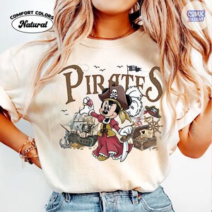 May include: Cream-colored t-shirt with a pirate-themed graphic. The design features a cartoon character dressed as a pirate, a pirate ship, treasure chest, and the word "Pirates". The shirt also has the words "Comfort Colors" and "Natural".