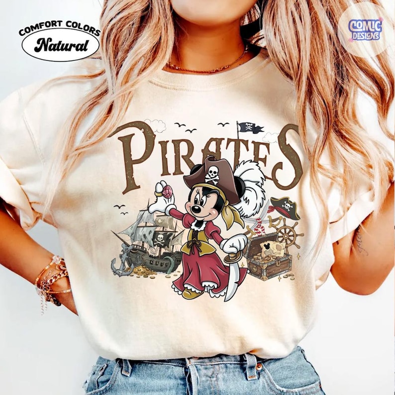 May include: Cream-colored t-shirt with a pirate-themed graphic. The design features a cartoon character dressed as a pirate, a pirate ship, treasure chest, and the word "Pirates". The shirt also has the words "Comfort Colors" and "Natural".