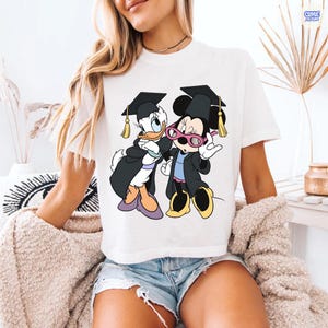 Puede incluir: Camiseta blanca con una ilustraci&oacute;n de dibujos animados de Daisy Duck y Minnie Mouse con atuendo de graduaci&oacute;n. Los personajes llevan birretes y togas de graduaci&oacute;n. Daisy Duck lleva zapatos morados y Minnie Mouse gafas rosas.