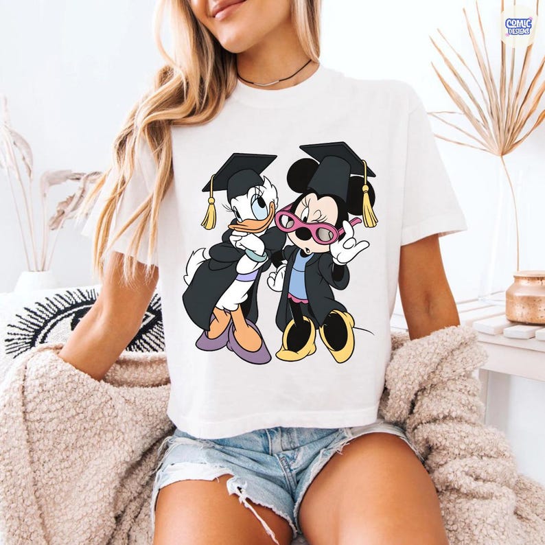 Puede incluir: Camiseta blanca con una ilustraci&oacute;n de dibujos animados de Daisy Duck y Minnie Mouse con atuendo de graduaci&oacute;n. Los personajes llevan birretes y togas de graduaci&oacute;n. Daisy Duck lleva zapatos morados y Minnie Mouse gafas rosas.