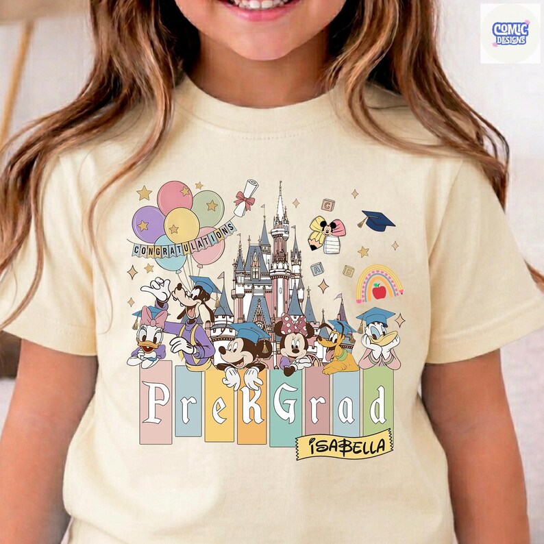 May include: A light yellow t-shirt featuring a colorful graphic of Disney characters, a castle, balloons, and the text "PreK Grad" with the name "Isabella". The design includes graduation caps and a rainbow.