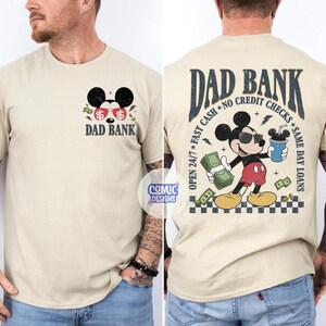 May include: Beige t-shirt with a graphic of Mickey Mouse wearing sunglasses and holding money and a drink. The front of the shirt says "DAD BANK" with dollar signs. The back of the shirt has the text "DAD BANK" and "OPEN 24/7 FAST CASH NO CREDIT CHECKS SAME DAY LOANS".