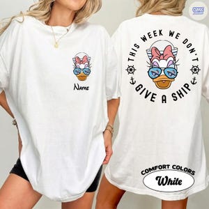 May include: White t-shirt featuring a cartoon duck wearing sunglasses and a bow. The front of the shirt has the word "Name" below the duck. The back of the shirt has the phrase "This week we don't give a ship" in a circular design.