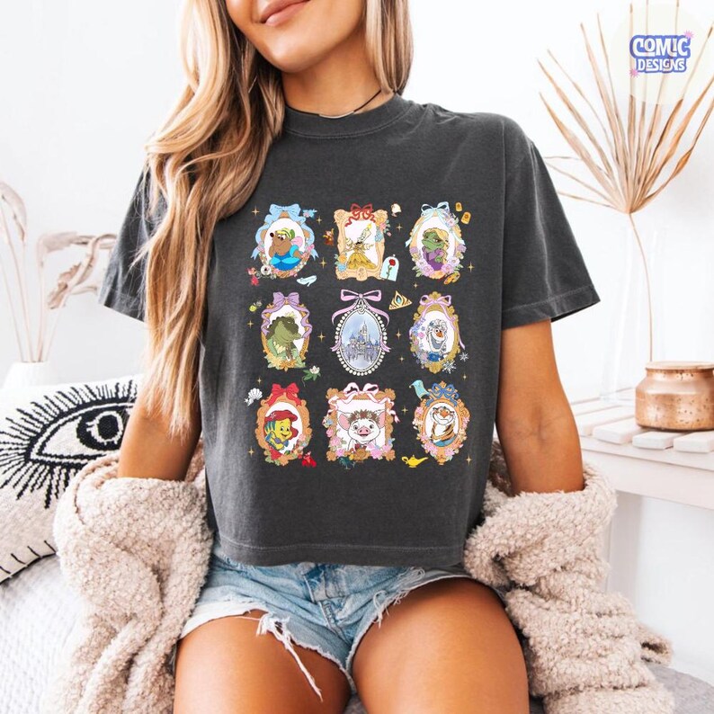 Puede incluir: Camiseta gris oscuro con nueve retratos enmarcados de personajes de dibujos animados. Cada retrato presenta un personaje diferente, incluyendo un oso, una princesa y un drag&oacute;n. La camiseta tiene las palabras "COMIC DESIGNS" en un recuadro azul.