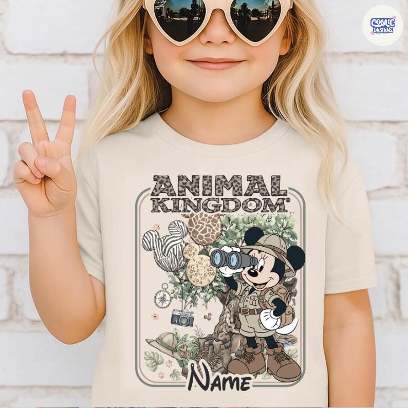 May include: A beige t-shirt featuring the words "ANIMAL KINGDOM" in a leopard print font. The shirt has a cartoon illustration of Minnie Mouse in safari attire, holding binoculars. The design includes animal print Mickey Mouse ears and a camera.