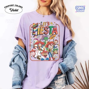 May include: Violet Comfort Colors t-shirt with a colorful "Let's Fiesta" graphic featuring cartoon characters in sombreros. The design includes musical notes and stars, with the Comic Designs logo in the upper right corner.