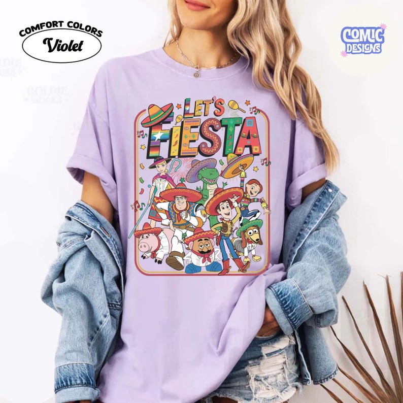 May include: Violet Comfort Colors t-shirt with a colorful "Let's Fiesta" graphic featuring cartoon characters in sombreros. The design includes musical notes and stars, with the Comic Designs logo in the upper right corner.