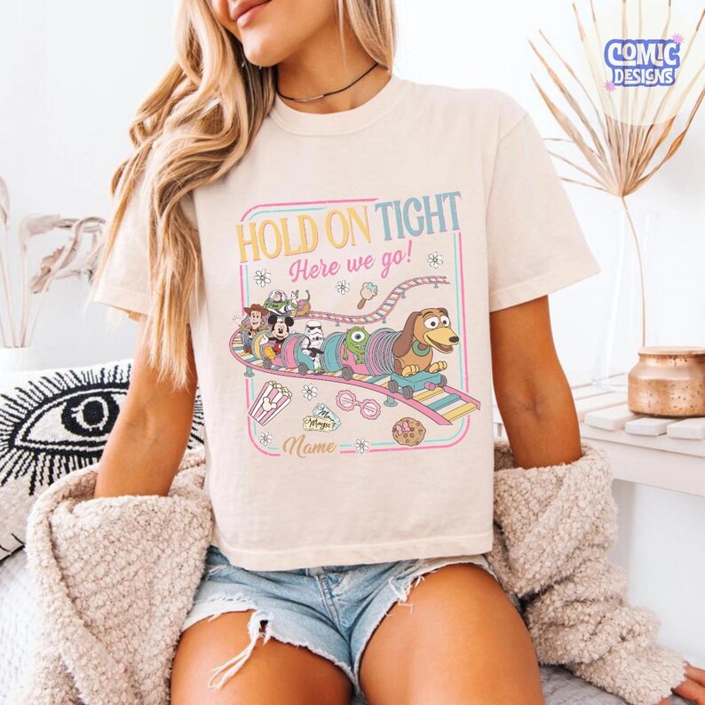 May include: Cream-colored t-shirt with a colorful graphic featuring cartoon characters on a roller coaster. The text "HOLD ON TIGHT Here we go!" is displayed above the characters. The design includes a space for a name.
