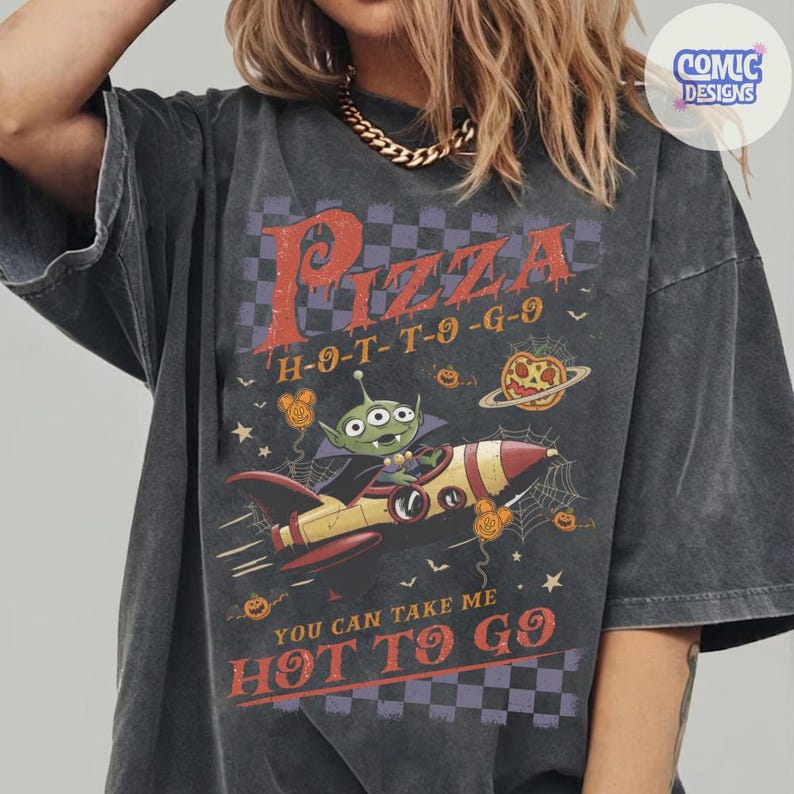 May include: A charcoal gray short-sleeve t-shirt featuring a graphic of a green three-eyed alien in a rocket ship. The design includes the text Pizza H-O-T-T-O-G-O and You Can Take Me Hot To Go, surrounded by spiderwebs, stars, and pumpkins.