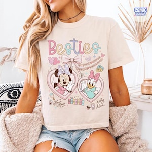 May include: Cream-colored t-shirt featuring the word "Besties" in pastel colors. The shirt showcases Minnie Mouse and Daisy Duck in heart-shaped lockets. Additional details include a bow, balloons, and the text "Minnie Mouse" and "Daisy Duck".