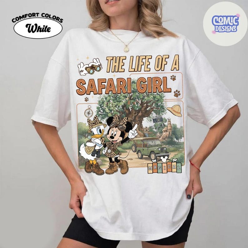 May include: White Comfort Colors t-shirt featuring a graphic of Minnie and Daisy Duck in safari outfits, with the text "THE LIFE OF A SAFARI GIRL." The design includes a jeep, tree, and the word "MAGICAL."