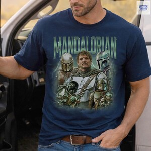 May include: Navy blue t-shirt featuring a graphic of characters from The Mandalorian. The design includes the word "MANDALORIAN" in green text above the characters. The image includes multiple characters from the show.