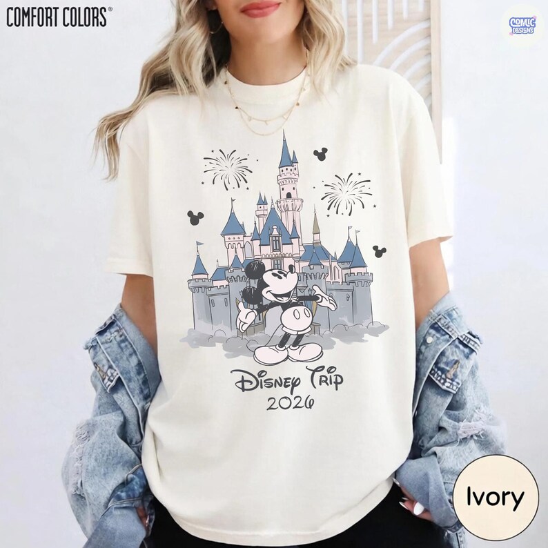 May include: Ivory-colored t-shirt featuring a graphic of Mickey Mouse in front of a castle with fireworks. The text "Disney Trip 2026" is below the graphic. The shirt is from Comfort Colors.