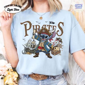 May include: Light blue t-shirt with a cartoon pirate design. The design features a blue alien character wearing a pirate hat and holding a sword and scroll. The word "PIRATES" is printed in brown letters. The shirt also has the words "COMFORT COLORS" and "Light Blue".