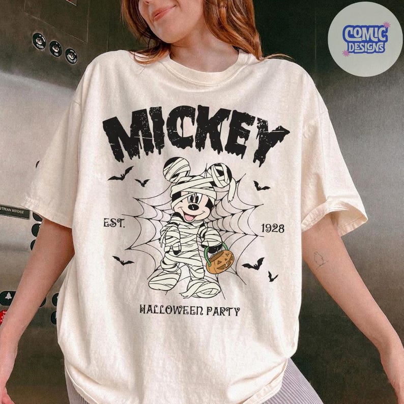 May include: A cream-colored short-sleeve t-shirt featuring a black graphic of Mickey Mouse wrapped as a mummy inside a spider web. The text reads MICKEY, EST. 1928, and HALLOWEEN PARTY. Small black bats are scattered around the central design.