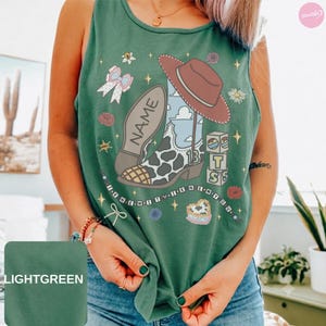 Puede incluir: Una camiseta sin mangas de color verde claro con un estampado gr&aacute;fico de una bota de vaquero con patr&oacute;n de manchas de vaca, un sombrero marr&oacute;n, un lazo y bloques. El texto "NAME" aparece en la bota y "I KNEW IT I KNEW YOU" debajo.