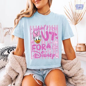 May include: Light blue t-shirt with a pink graphic featuring Daisy Duck, the text "school's out for Disney", and various Disney-themed icons. The shirt has short sleeves and a crew neck.