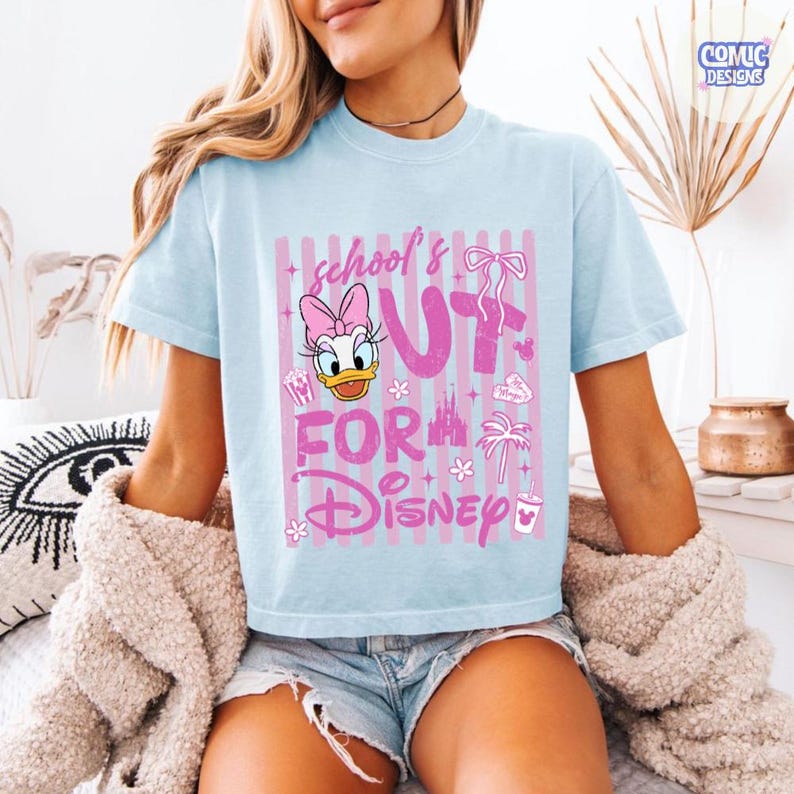 May include: Light blue t-shirt with a pink graphic featuring Daisy Duck, the text "school's out for Disney", and various Disney-themed icons. The shirt has short sleeves and a crew neck.